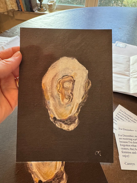 Hand holding a small art print of an oyster against a black background against a kitchen table