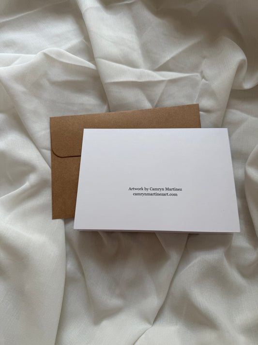 The back of a greeting card with text that reads "Artwork by Camryn Martinez camrynmartinezart.com" against an envelope with a white fabric background