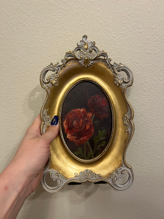 Decorative gold frame with a painting of red ranunculus against a dark green background. The painting is being held by a hand.