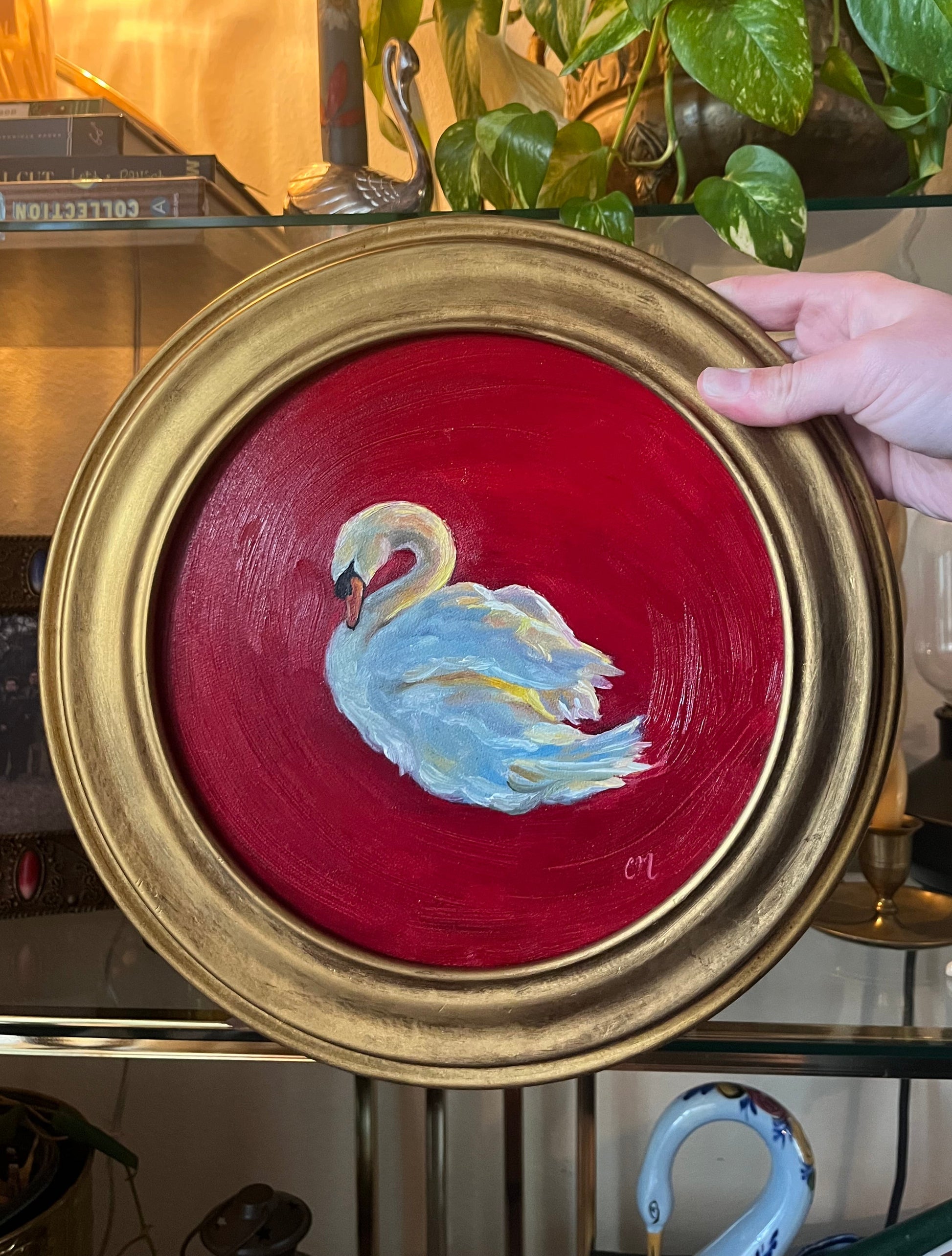 Round oil painting with a swan design on a red background and gold frame held by a hand.