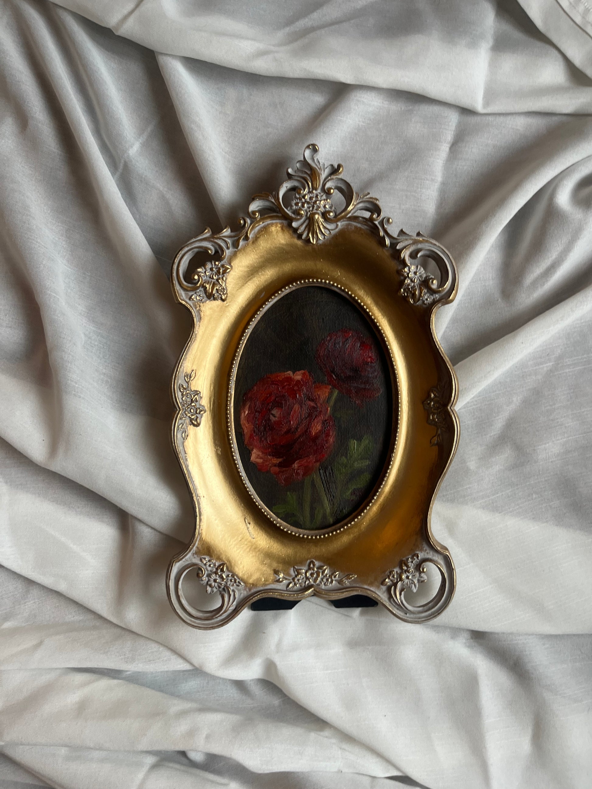 Decorative gold frame with a painting of red ranunculus against a dark green background. The painting is against a white fabric background.