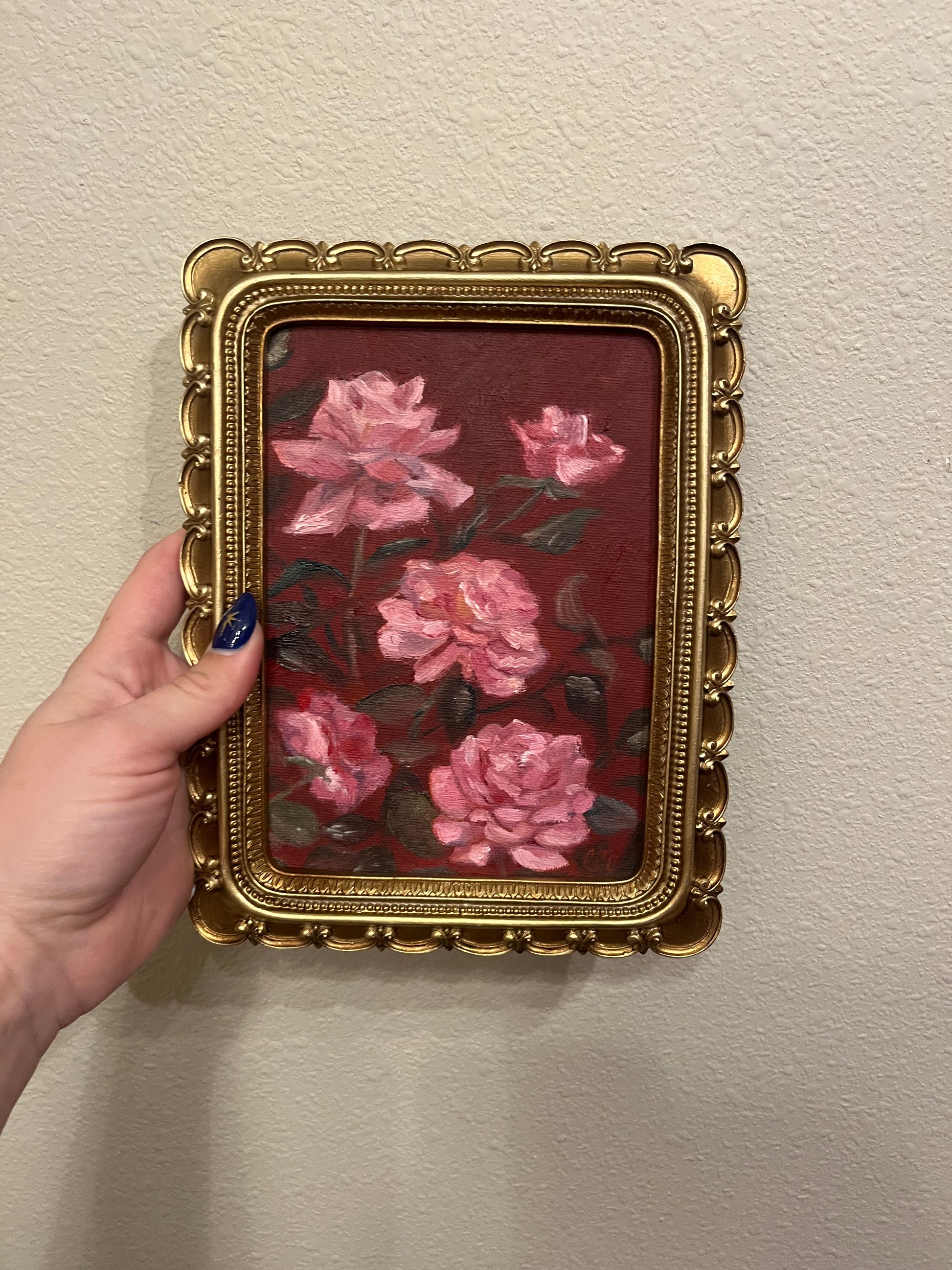 Decorative painting of pink flowers against a dark red background in a gold frame held by a hand against a beige wall.