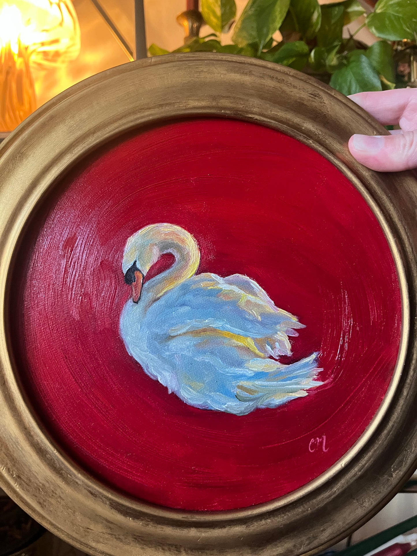 Round oil painting with a swan design on a red background and gold frame held by a hand.
