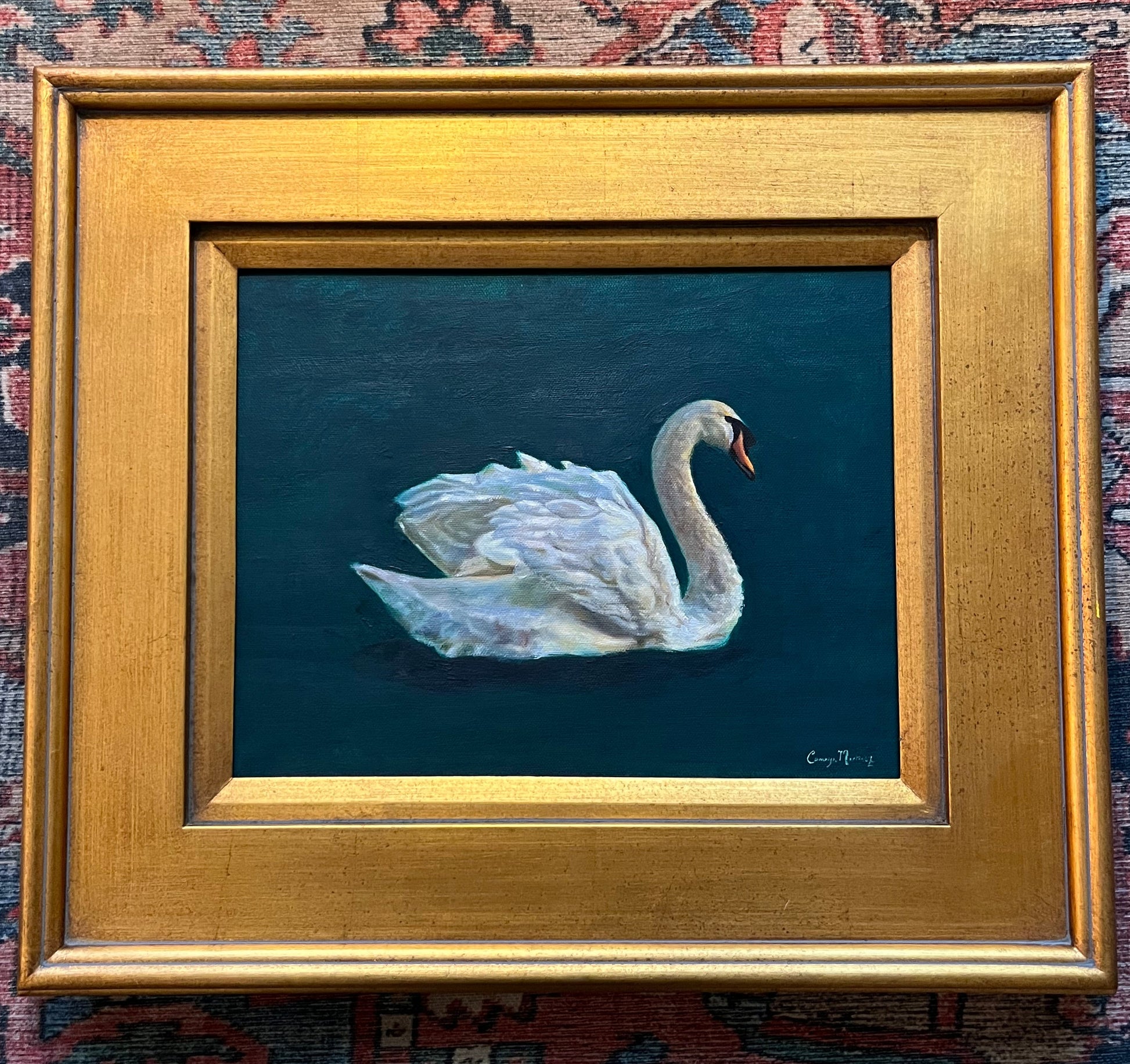 Framed artwork of a swan on a dark green background with a gold plein air frame displayed on a patterned fabric background.