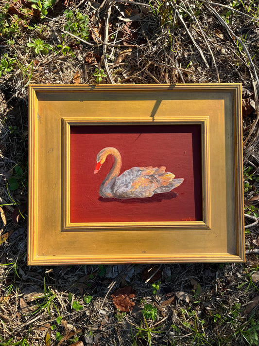 Framed artwork of a swan on a red background and gold plein air frame placed on dry grass.